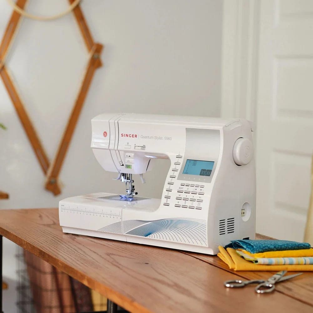 SINGER Quantum Stylist 9960 Nähmaschine auf einem Holztisch, daneben Stoffe und eine Schere